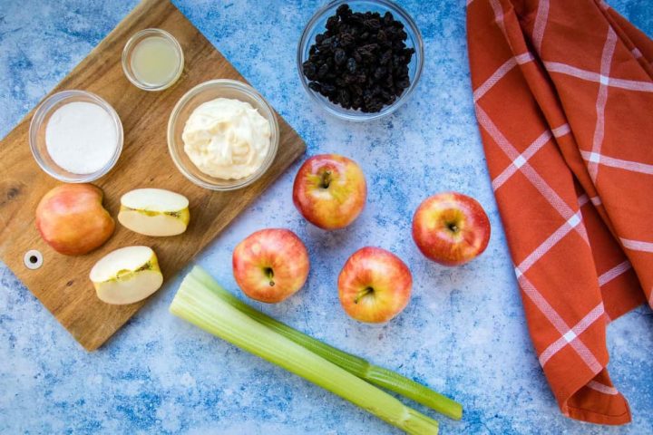 Overhead view of 4 5 apples (one chopped), two stalks of celery, a bowl of raisins, a bowl with mayonnaise, a small bowl with lemon juice, and a small bowl with sugars all on a blue surface with a coral plaid towel to the side