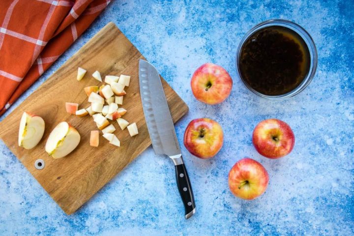 Overhead view of a chopping board with shopped apple and a knife, four apples to the side, a bowl of raisins soaking in water, all on a blue surface with a coral towel laying to the side.