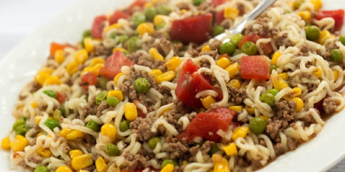 Ground beef, corn, peas, diced tomotoes, and ramen noodles on a white plate.