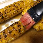 Grilled corn in a clear Pyrex dish with a silicone brush basting with garlic ginger soy butter