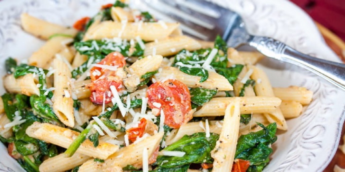 White plate with penne pasta, cooked baby spinach, cherry tomatoes, and shredded mozzarella cheese. There is a fork on the plate.