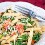 White plate with penne pasta, cooked baby spinach, cherry tomatoes, and shredded mozzarella cheese. There is a fork on the plate.