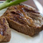 A cooked steak on a white plate with a stalk of celery in the background.