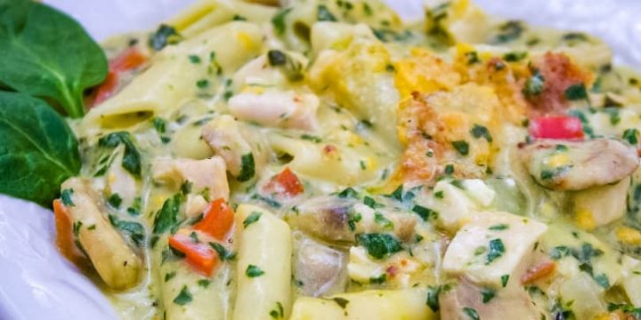 Penne pasta with mushrooms, diced red bell pepper, chopped spinach, and cubed chicken in a cream of chicken sauce, topped with shredded cheddar cheese and garnished with two baby spinach leaves on a white plate.