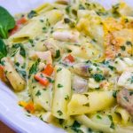 Penne pasta with mushrooms, diced red bell pepper, chopped spinach, and cubed chicken in a cream of chicken sauce, topped with shredded cheddar cheese and garnished with two baby spinach leaves on a white plate.