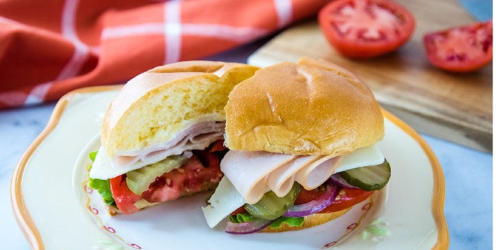 Brioche bun with slices of turkey, Swiss cheese, lettuce, tomato, pickle, and onion on a white and orange plate with cutting board and tomato in the background