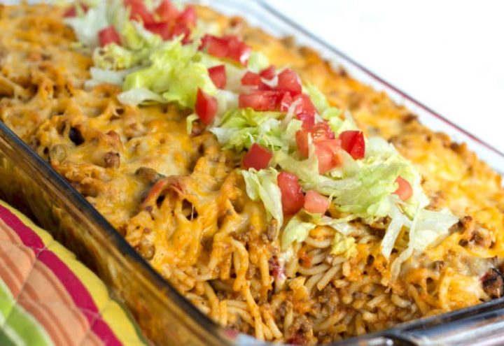Clear casserole dish with spaghetti noodles, ground beef, and melted cheese, topped with shredded lettuce and diced tomatoes.