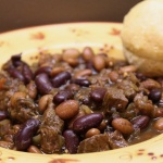 Kidney beans, pinto beans, and diced beef in a broth, all in a yellow soup bowl with red flowers. There is a biscuit on the rim of the bowl.