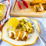 Photo is a clear glass bowl of diced potatoes sitting on an orange and white striped linen napkinn, a wooden cutting board with tongs and sliced chicken, and a plate with a small corn cob, diced potatoes, and sliced chicken. The plate is off-white with a yellow rim. There is a fork on the plate beside the potatoes.