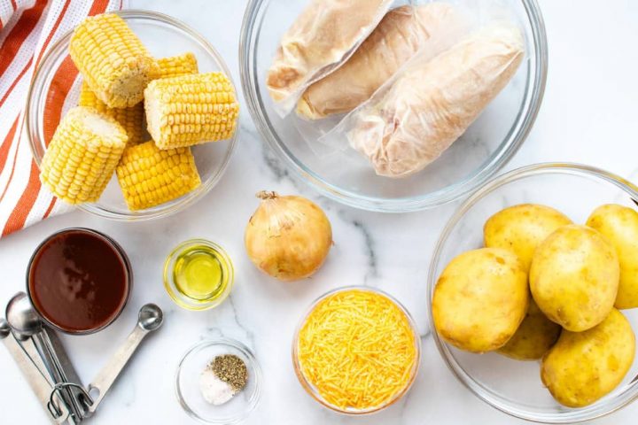 Clear glass bowl with small corn on the cob, clear glass bowl with individually wrapped chicken breasts, clear glass bowl of whole, unpeeled potatoes, bowl of shredded cheddar cheese, bowl with salt and pepper, metal measuring spoons, bowl of barbecue sauce, bowl of oil, and an onion, all on a white marble countertop with an orange and white striped napkin.