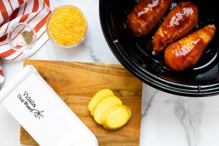 Small bowl with salt and pepper sitting on an orange and white striped napkin, a bowl of shredded cheddar cheese, a slow cooker with three chicken breasts in barbecue sauce, a wooden cutting board with sliced potatoes, and a Vidalia Chop Wizard.