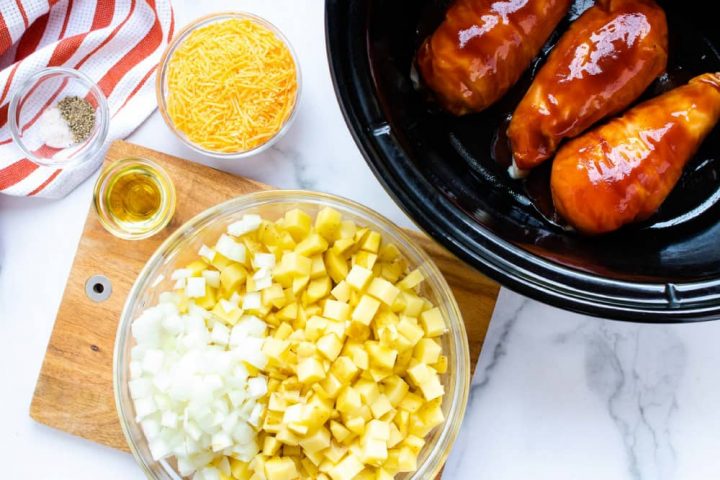 Small bowl with salt and pepper sitting on an orange and white striped napkin, a bowl of shredded cheddar cheese, a slow cooker with three chicken breasts in barbecue sauce, a wooden cutting board a bowl of diced potatoes and onion, and a small bowl with oil.