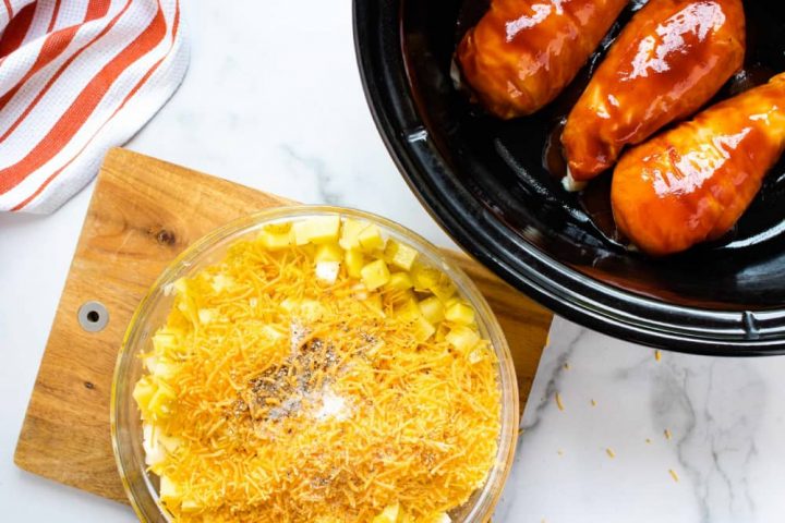 An orange and white striped linen napkin on a white marble countertop with a slow cooker with three chicken breasts in barbecue sauce. A wooden cutting board with a bowl of diced potatoes and onions, shredded cheddar cheese, and salt and pepper.