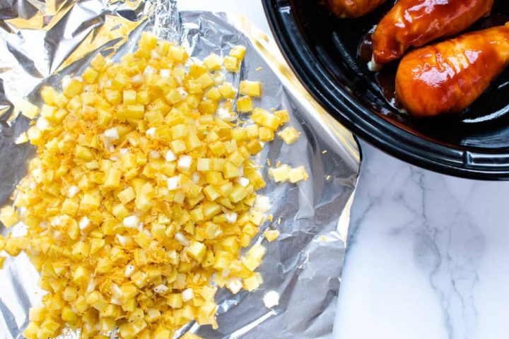 A large piece of foil with diced potatoes, shredded cheddar cheese, and diced onions, beside a slow cooker with chicken breasts in barbecue sauce.