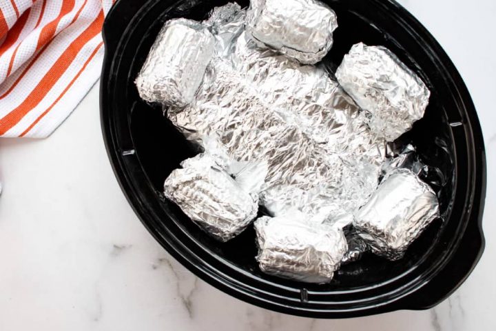 Slow cooker with six small corn cobs wrapped in foil and another foil packet beneath them.
