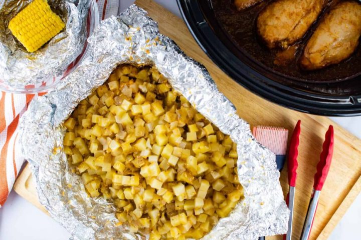 A bowl with foil and a small corn cob, open foil packet with diced potatoes, a slow cooker with chicken breasts. The potatoes are on a wooden cutting board with a basting brush and a set of tongs.