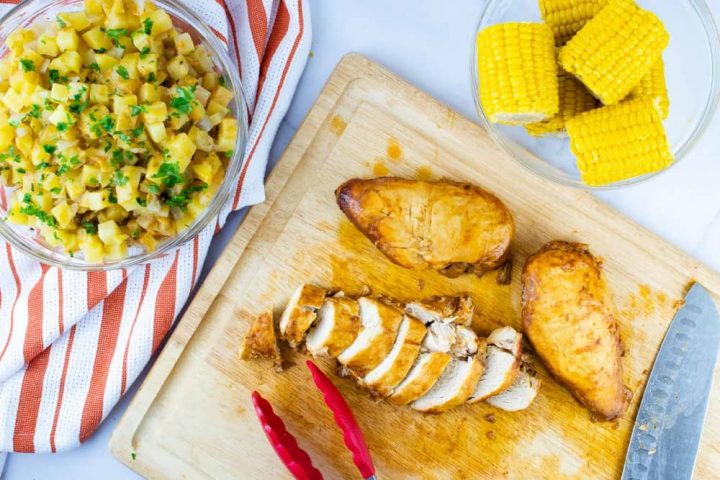 A clear glass bowl with diced potatoes sitting on an orange and white striped napkin, a clear glass bowl with small corn cobs, and a wooden cutting board with two whole chicken breasts and one sliced chicken breast. There is a set of tongs and a Santoku Knife on the cutting board.