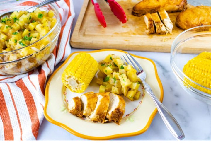 Photo is a clear glass bowl of diced potatoes sitting on an orange and white striped linen napkinn, a wooden cutting board with tongs and sliced chicken, a clear glass bowl of corn on the cob, and a plate with a small corn cob, diced potatoes, and sliced chicken. The plate is off-white with a yellow rim. There is a fork on the plate beside the potatoes.