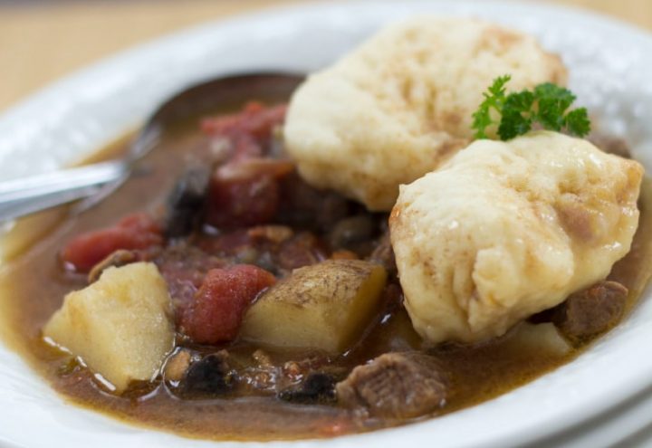 White bowl of beef stew with two dumplings on top. Stew consists of brown broth, diced beef, chopped potatoes, and stewed tomatoes. There is a spoon in the bowl on the left side.