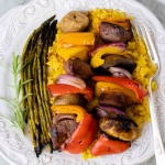 grilled kabobs of red, yellow, and orange bell pepper pieces alternated with mushrooms, red onion pieces, and marinated beef and layered over yellow rice with grilled asparagus on a white plate with a silver fork