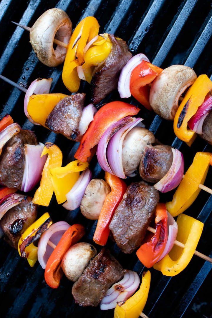kabobs of red, yellow, and orange bell pepper pieces alternated with mushrooms, red onion pieces, and marinated beef on a grill