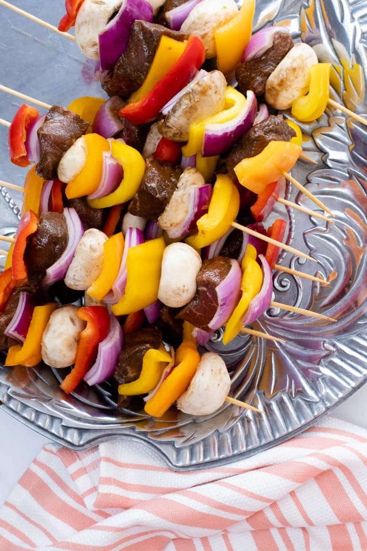 uncooked kabobs of beef alternated with red, yellow, and orange bell peppers, mushrooms, and red onions on a pewter platter