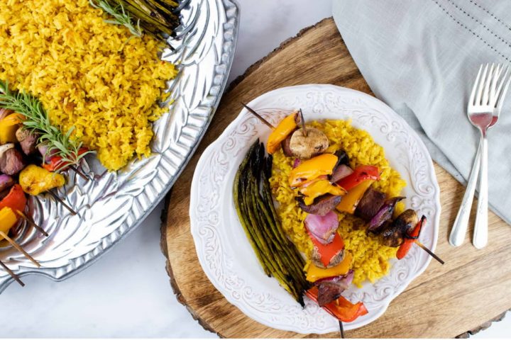 grilled kabobs of red, yellow, and orange bell pepper pieces alternated with mushrooms, red onion pieces, and marinated beef and layered over yellow rice with grilled asparagus on a white plate with a silver fork with a serving platter of kabobs, yellow rice, and asparagus to the side