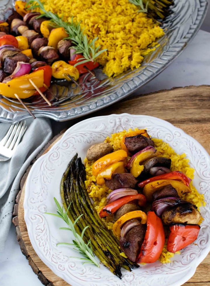 grilled kabobs of red, yellow, and orange bell pepper pieces alternated with mushrooms, red onion pieces, and marinated beef and layered over yellow rice with grilled asparagus on a white plate with a silver fork with a serving platter of kabobs, yellow rice, and asparagus to the side