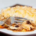 A tortilla covered with melted cheese, on top of ground beef, black beans, and tomatoes, all on a white plate. A fork is cutting into the tortilla.