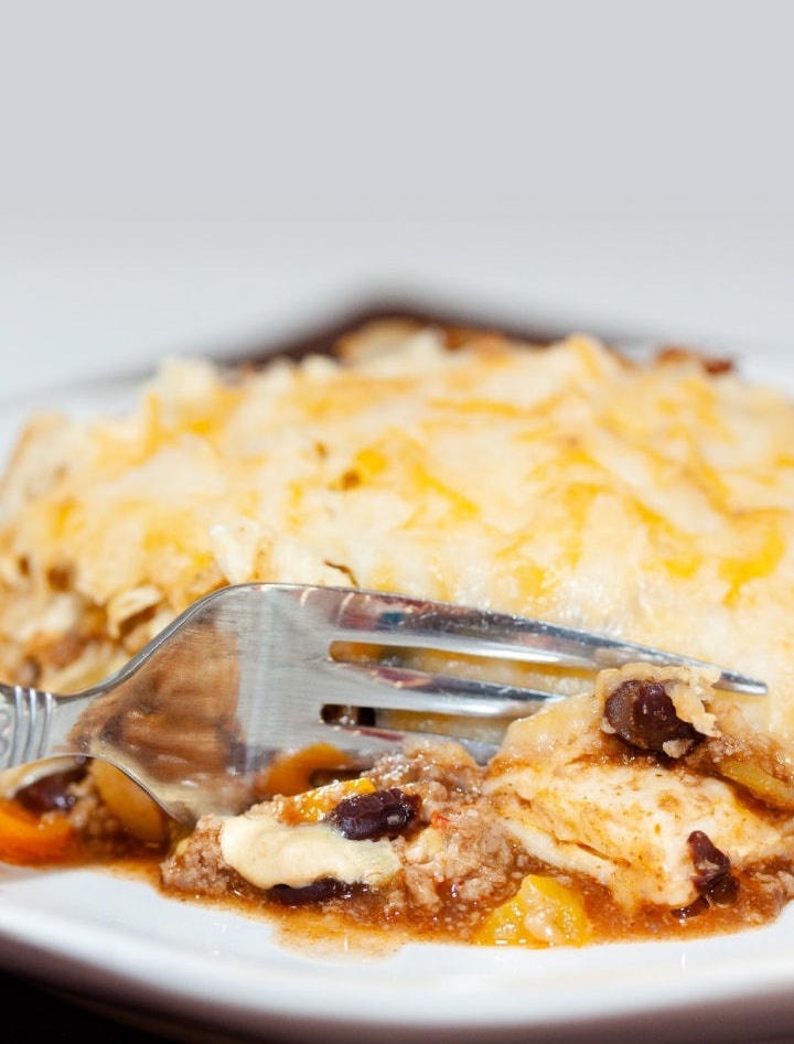 A tortilla covered with melted cheese, on top of ground beef, black beans, and tomatoes, all on a white plate. A fork is cutting into the tortilla.