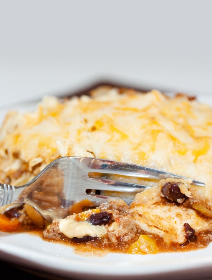 A tortilla covered with melted cheese, on top of ground beef, black beans, and tomatoes, all on a white plate. A fork is cutting into the tortilla.