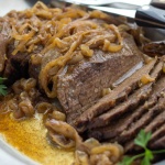 Slices of beef on a white platter. Beef is covered with cooked onions and resting in an au jus. The plate is garnished with parsley and there is a steak knife resting on the back of the platter.