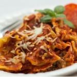 Ground beef, lasagna noodles, and melted mozarella cheese in a white bowl, with pepperoni on the right side and a fork on the left.