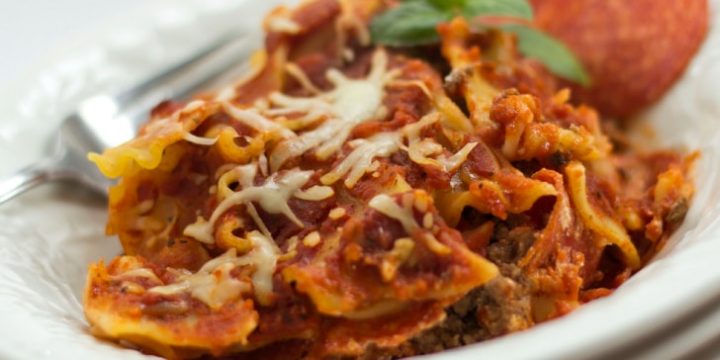 Ground beef, lasagna noodles, and melted mozarella cheese in a white bowl, with pepperoni on the right side and a fork on the left.