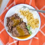 a white plate half full of roast beef and half full of rice. Roast beef and rice are both covered with gravy. There is a fork on the plate next to the rice. The plate is on an orange napkin with white stripes.