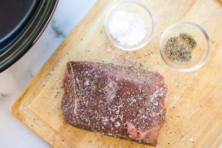 An uncooked beef roast sprinkled with salt and pepper on a wooden cutting board next to a slow cooker. There is a small glass bowl of salt and a small glass bowl of pepper on the cutting board next to the roast.