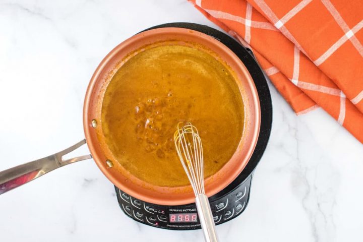 Overhead shot of a small orange skillet of brown gravy over a warming unit on a white marble counter.There is a whisk in the skillet, and an orange napkin with white stripes on the counter at the top right of the picture.