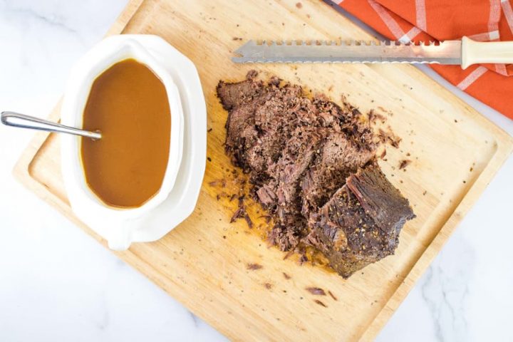 White gravy bowl on a white platter. The bowl has brown gravy and a spoon. The platter is sitting on a wooden cutting board next to a beef roast that has been half-sliced with the serrated knife at the top of the photo. There is an orange napkin with white stripes in the top right corner of the photo.