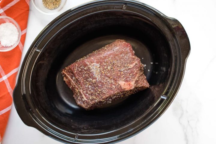 Uncooked beef roast in a slow cooker on a white marble counter. On the left side of the photo is an orange napkin with white stripes. On the napkin is a small glass bowl with salt. Next to the napkin is small glass bowl with pepper.