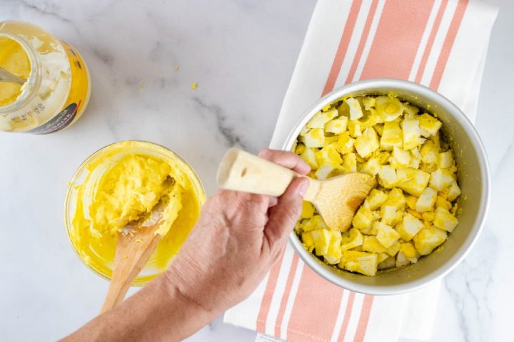 Overhead view of hard boiled eggs being broken up with a wooden spatula with a separate bowl containing a mayonnaise and yolk mixture