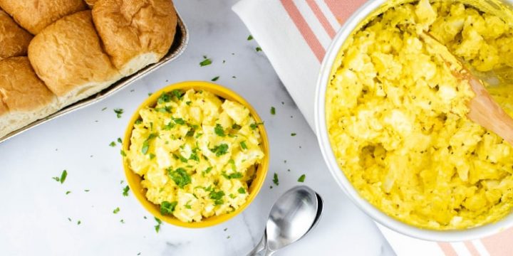 Prepared egg salad garnished with chopped parsley in a white bowl with a yellow rim with whole wheat rolls in the background