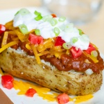 Baked potato topped with chili, shredded cheddar cheese, diced tomatoes, sour cream, and diced green onions, with shredded cheese and diced tomatoes are on the counter around the potato.