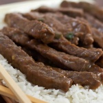 Sliced steak with a sauce on a bed of rice with chopsticks in the foreground.