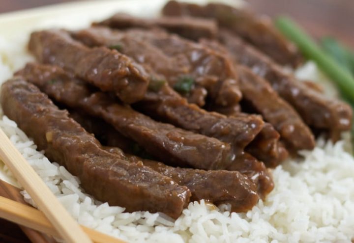 Sliced steak with a sauce on a bed of rice with scallions in the background and chopsticks in the foreground.