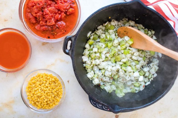 A white marble counter with a clear glass bowl of ditalini pasta, clear glass bowl of tomato sauce, clear glass bowl of fire-roasted tomatoes in tomato juice, and a cast iron stew pot with chopped celery and onions with Italian seasoning. The celery and onions are being stirred with a wooden spoon.