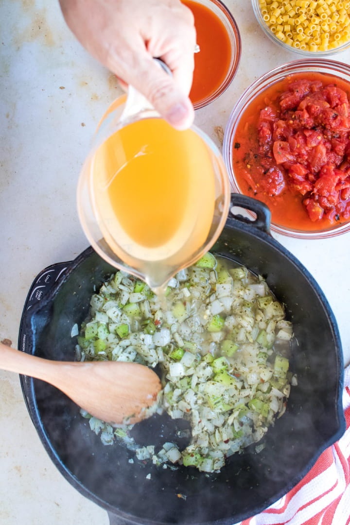 A measuring cup of broth is being poured into a cast iron stew pot of chopped celery and onions. A wooden spoon is stirring the contents of the pan. At the top right of the photo is a clear glass bowl with roasted tomatoes in tomato juice.