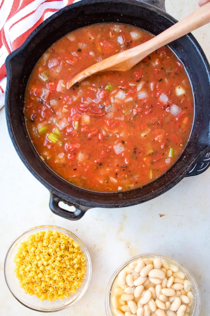 A cast iron stew pot with soup consisting of chopped celery and onions, tomatoes, chicken broth, and tomato sauce. There's a wooden spoon in the soup. There's a clear glass bowl of ditalini pasta and a clear glass bowl of cannellini beans at the bottom of the photo.