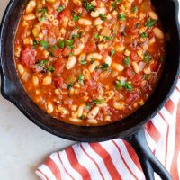 Cast iron skillet with Pasta e Fagioli, a soup made with pasta, tomatoes, chopped celery, onion, and parsley, cannellini beans, and tomato sauce. There's an orange and white striped towel on the counter to the right of the skillet. Text pasta e fagoioli (pasta & beans) add salt & serve formerly menus4moms