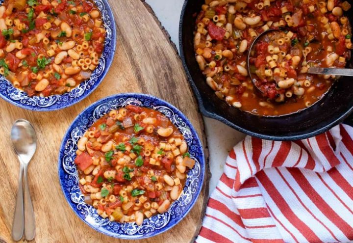 Pasta e Fagioli The left of the photo is two blue and white soup bowls, each filled with Pasta e Fagioli, a soup made with pasta,tomatoes, chopped celery, onion, and parsley, cannellini beans, and tomato sauce. There are two spoons next to the bowls. On the right side of the bowls is a cast iron skillet with Pasta e Fagioli. There's an orange and white striped towel on the counter to the right of the skillet, and a ladle scooping soup out of the skillet.