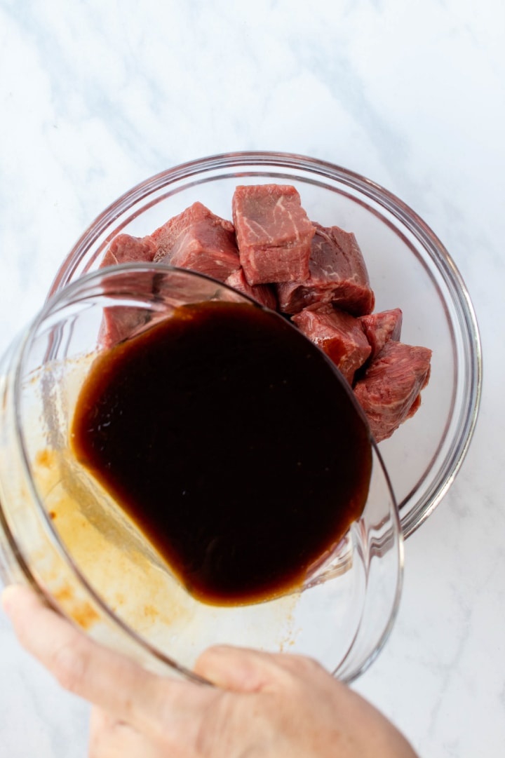 Large bowl of soy marinade being poured over another bowl full of cubes of beef